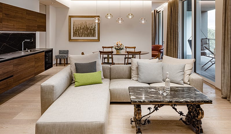 Lake View Family Grand Loggia Suite living room with a neutral L-shaped sofa, dark wood kitchen cabinetry, marble top coffee table, oval dining table, and glass doors to a private terrace at ARIA Retreat & SPA by Kenwood Travel