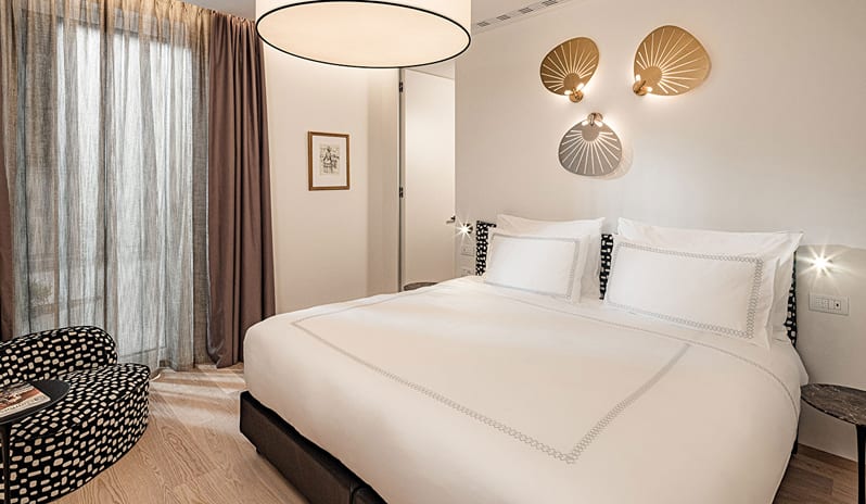 Lake View Family Grand Loggia Suite bedroom with white linen bedding, modern brass leaf wall sconces, black and white printed accent chair, and sheer curtains at ARIA Retreat & SPA by Kenwood Travel