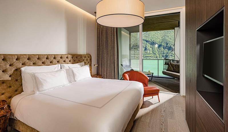 Lake View Family Grand Loggia Suite bedroom with a king bed, tufted headboard, leather accent chair, and sliding doors to a private lake-view balcony with a hanging swing lounge at ARIA Retreat & SPA by Kenwood Travel