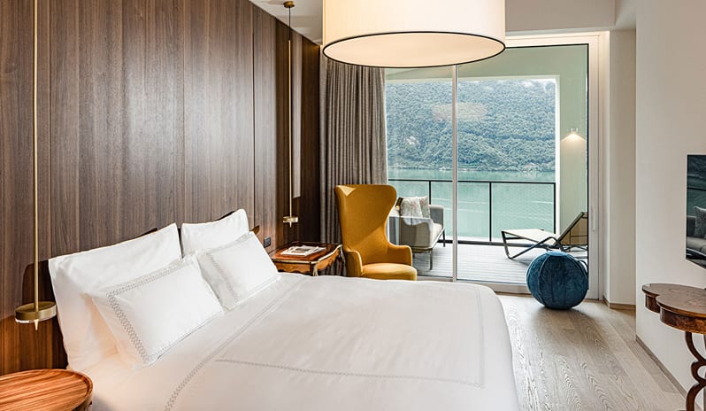 ARIA Retreat & SPA Lake View Grand Loggia Suite bedroom with a king bed, warm wood paneling, mustard yellow wingback chair, and large sliding glass doors leading to a private lake-view loggia terrace-Kenwood Travel