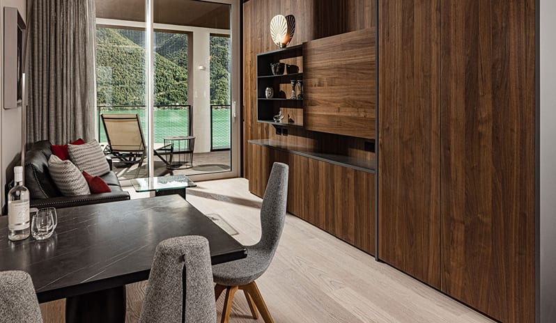 Lake View One Bedroom Loggia Suite living space with a round marble top dining table, grey upholstered chairs, rich dark wood storage cabinets, and floor-to-ceiling glass doors opening to a scenic lake terrace at & SPA by Kenwood Travel