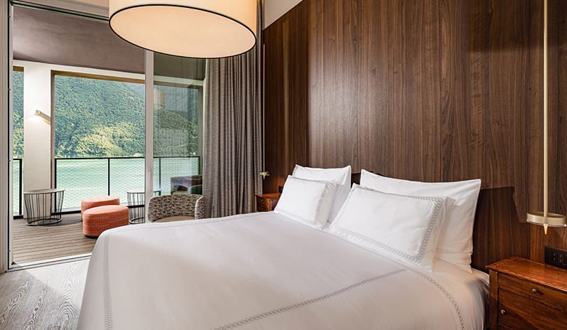 Lake View One Bedroom Loggia Suite king bed with dark wood accent wall, drum pendant lamp, and open sliding glass doors leading to a private balcony at ARIA Retreat & SPA by Kenwood Travel