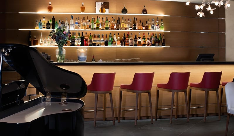 San Marco Lounge & Bar Modern lounge bar featuring red bar stools, a backlit liquor display, and a black grand piano at ARIA Retreat & SPA by Kenwood Travel