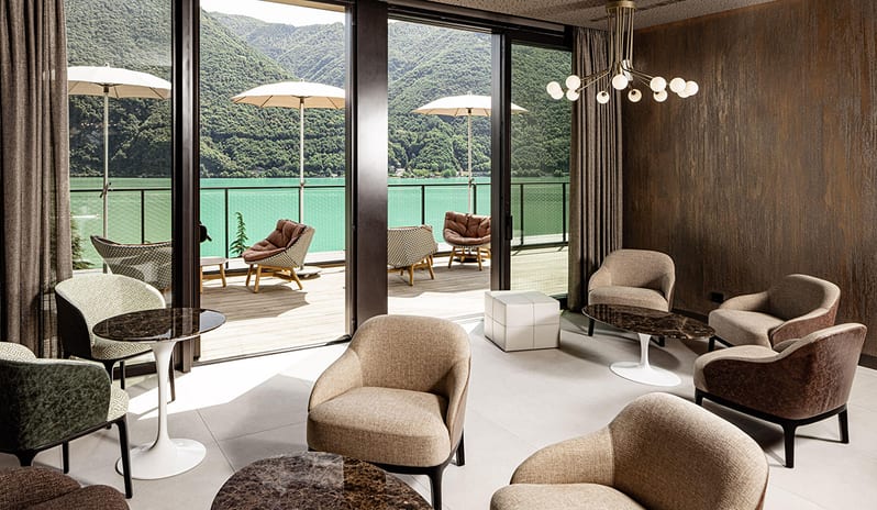 Dolce Vita Lounge & Terrace Contemporary lounge interior with textured accent walls, modern brass chandelier, and neutral armchairs facing an outdoor patio with lake views at ARIA Retreat & SPA by Kenwood Travel