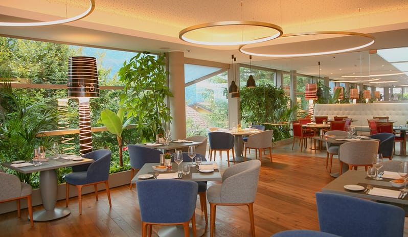 La Joie de Vivre Restaurant Spacious luxury hotel dining room with elegant table settings, indoor tropical plants, and panoramic scenic window views at ARIA Retreat & SPA by Kenwood Travel