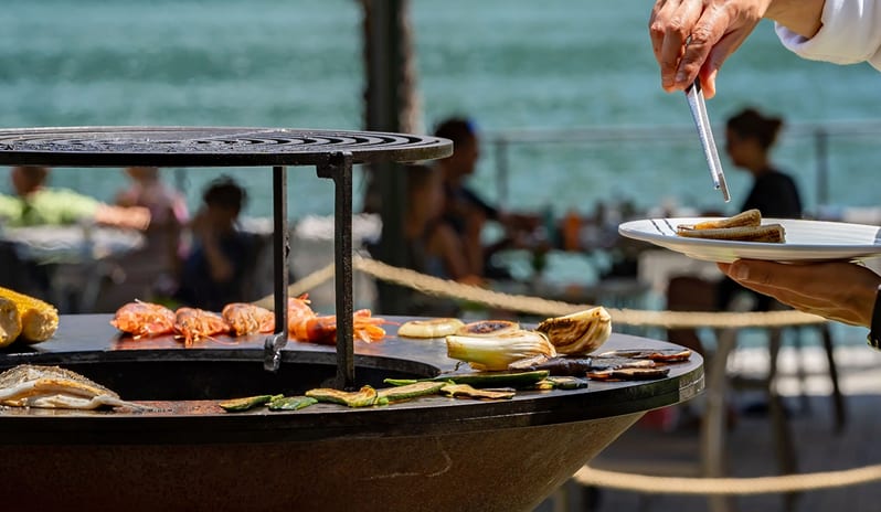 46° Parallelo A chef serving fresh barbecue seafood on a plate at a scenic waterfront outdoor restaurant at ARIA Retreat & SPA by Kenwood Travel