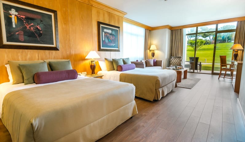 Family Suite two double beds with beige and purple bedding, wood paneling walls, and glass doors leading to a green lawn at Gloria Verde by Kenwood Travel