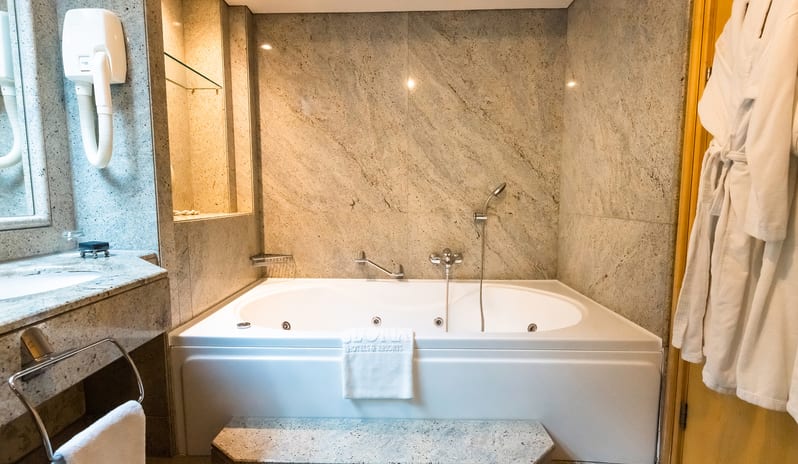 Family Suite bathroom showcasing a deep jetted soaking tub, granite countertops, a wall-mounted hair dryer, and a plush white bathrobe hanging on the door at Gloria Verde by Kenwood Travel