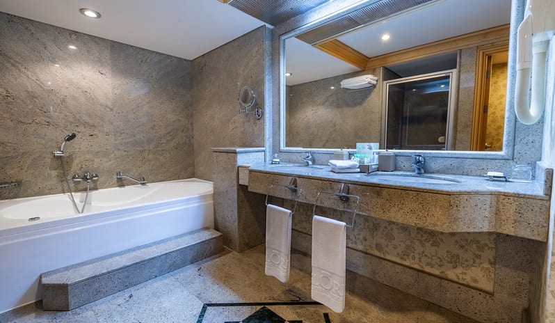 Family Suite bathroom showing a double washbasin setup, hotel toiletries, a wall-mounted magnifying mirror, and patterned floor tile details at Gloria Verde by Kenwood Travel