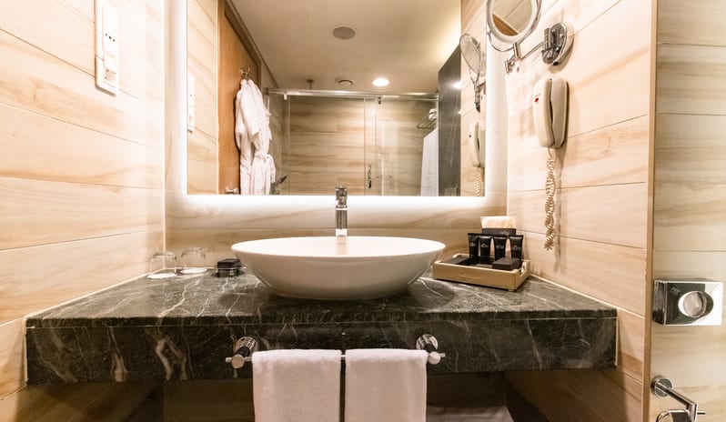 Family Suite bathroom showing a modern washbasin setup, wood-look tiled walls, a magnifying mirror, and a reflection of the walk-in shower at Gloria Verde by Kenwood Travel