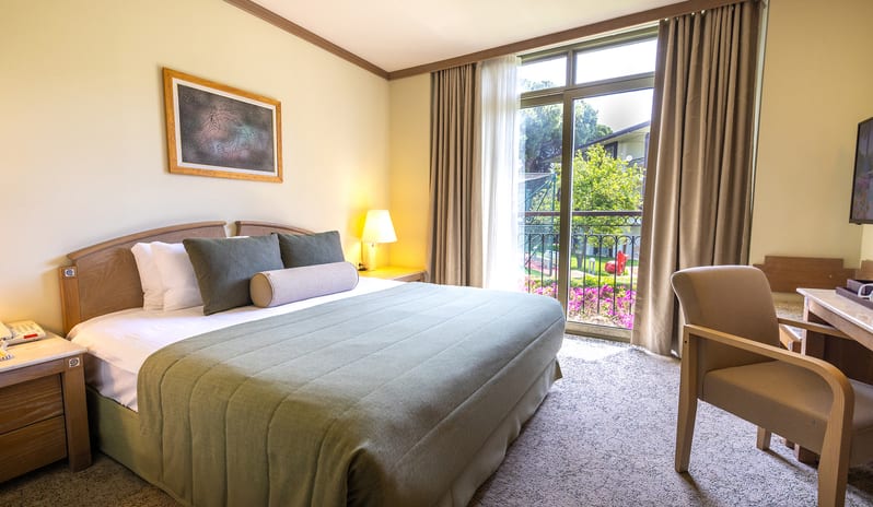 Family Suite large bed with sage green bedding, an armchair at a writing desk, and floor-to-ceiling glass doors opening to a garden view at Gloria Verde by Kenwood Travel