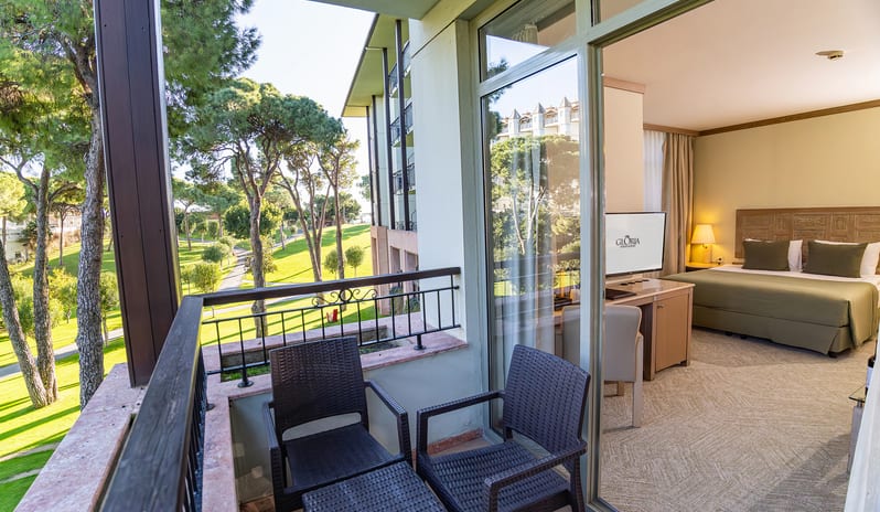 Standard Room balcony with outdoor seating, glass sliding doors, and a view of the hotel room interior and surrounding green grounds at Gloria Verde by Kenwood Travel