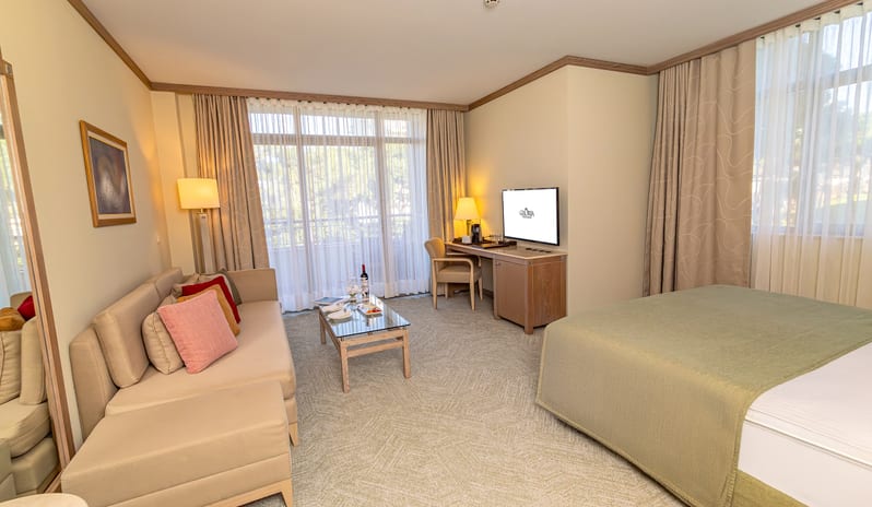 Standard Room seating area with a beige sofa, a glass coffee table, a desk with a TV, and a double bed at Gloria Verde by Kenwood Travel