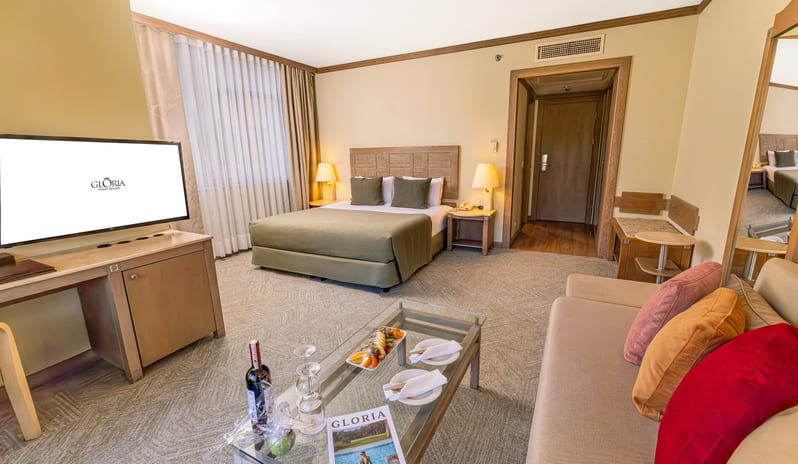 Standard Room foreground seating, a media console, a large bed with green accents, and an open doorway leading to the hallway at Gloria Verde by Kenwood Travel