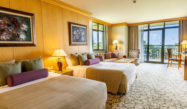 King Suite two large beds with olive and purple bolster pillows, wood panel walls, and a balcony overlooking the resort at Gloria Verde by Kenwood Travel
