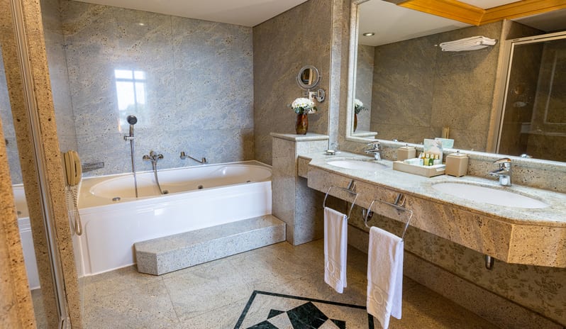 King Suite bathroom showcasing two washbasins, upscale hotel toiletries, a wall-mounted magnifying mirror, and a whirlpool bathtub setup at Gloria Verde by Kenwood Travel