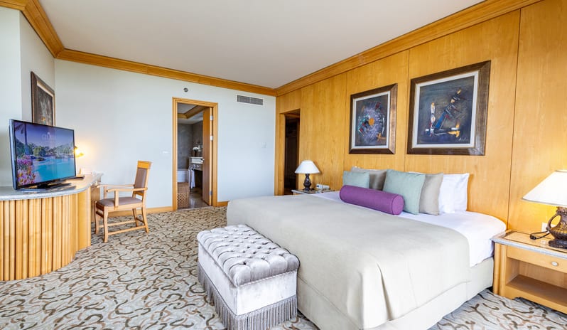 King Suite bedroom interior showcasing patterned carpeting, a TV console with a desk chair, framed wall art, and elegant bedside lamps at Gloria Verde by Kenwood Travel