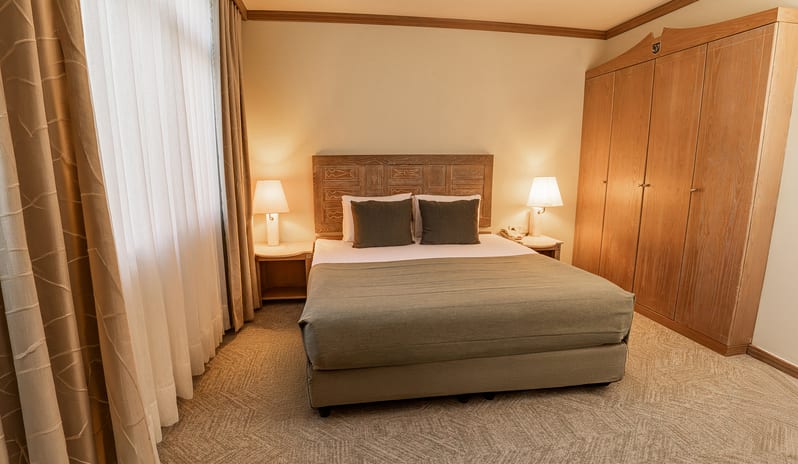 Suite double bed with olive green bedding, a large wooden wardrobe, and bedside lamps next to a window with sheer curtains at Gloria Verde by Kenwood Travel