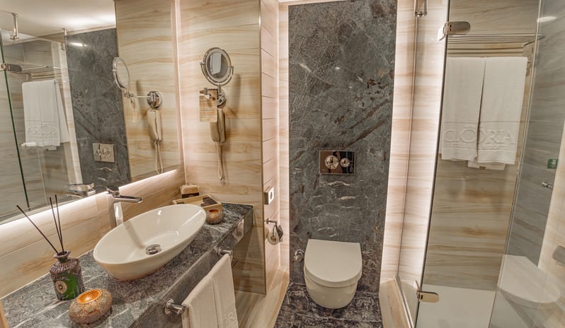 Suite white oval vessel sink on a dark grey marble vanity, a glass walk-in shower, a toilet, and large mirrors at Gloria Verde by Kenwood Travel
