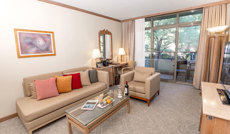 Suite living space showcasing a couch setup, a small writing desk with a mirror, a media console, and a view of the outside trees through large windows at Gloria Verde by Kenwood Travel