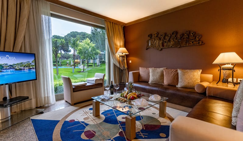 Select Villa brown leather sofa, a glass coffee table with fresh fruit, a colorful geometric rug, and a large window overlooking the resort pool and garden at Gloria Verde by Kenwood Travel