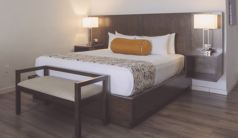 Hillside Suite Garden/Resort Views king bed with an extended dark wood headboard, warm bedside lamps, an orange bolster pillow, and a bed-end bench at The Verandah Antigua by Kenwood Travel