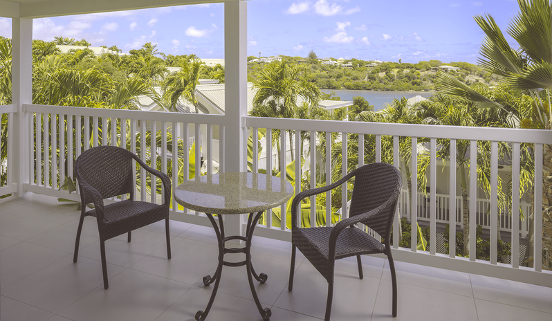 Water View Suite balcony with table and chairs overlooking lush palm trees and water at The Verandah Antigua by Kenwood Travel
