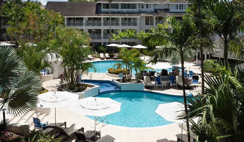 Garden View swimming pool deck with lounge chairs, white sun umbrellas, and palm trees at The Club Barbados Resort & Spa-Garden View by Kenwood Travel