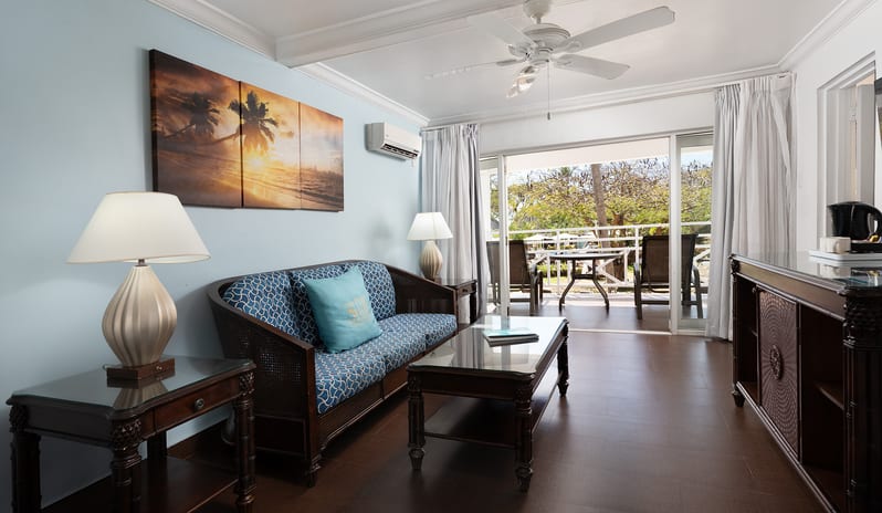 One Bedroom Pool Garden View Suite living area with a blue couch, coffee table, and balcony access at The Club Barbados Resort & Spa by Kenwood Travel