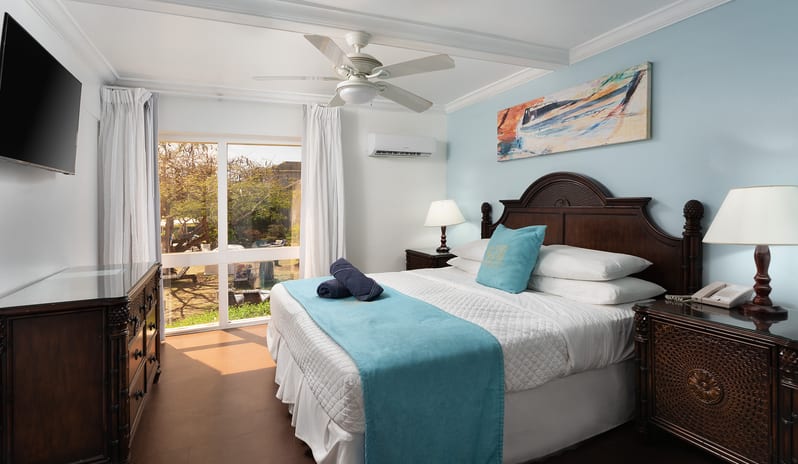 One Bedroom Pool Garden View Suite bedroom with dark wood nightstands and large window with garden view at The Club Barbados Resort & Spa by Kenwood Travel