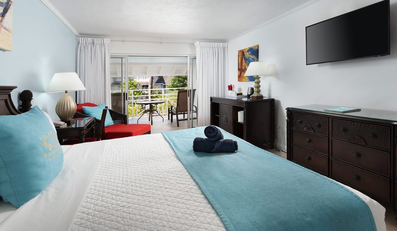Superior Garden View suite with dark wood dresser, mounted TV, and open balcony door at The Club Barbados Resort & Spa by Kenwood Travel