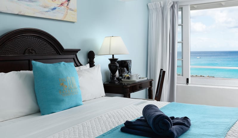 Ocean Loft Ocean View Hotel room interior featuring a dark wood dresser, TV, and a view of the ocean through the window at The Club Barbados Resort & Spa by Kenwood Travel