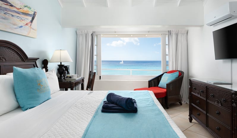 The Club Barbados Resort & Spa Ocean Loft Ocean View Hotel room interior featuring a dark wood dresser, TV, and a view of the ocean through the window-Kenwood Travel