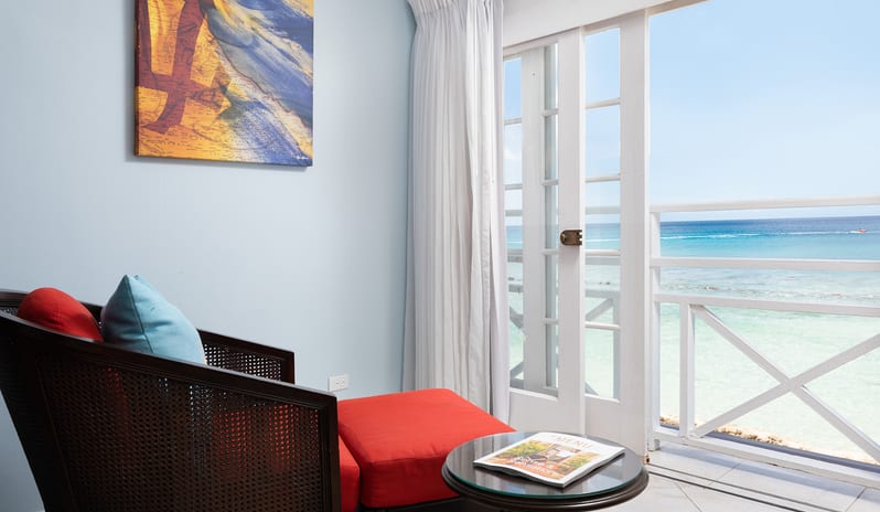 Superior Garden View armchair and ottoman next to an open balcony door with an ocean view at The Club Barbados Resort & Spa by Kenwood Travel