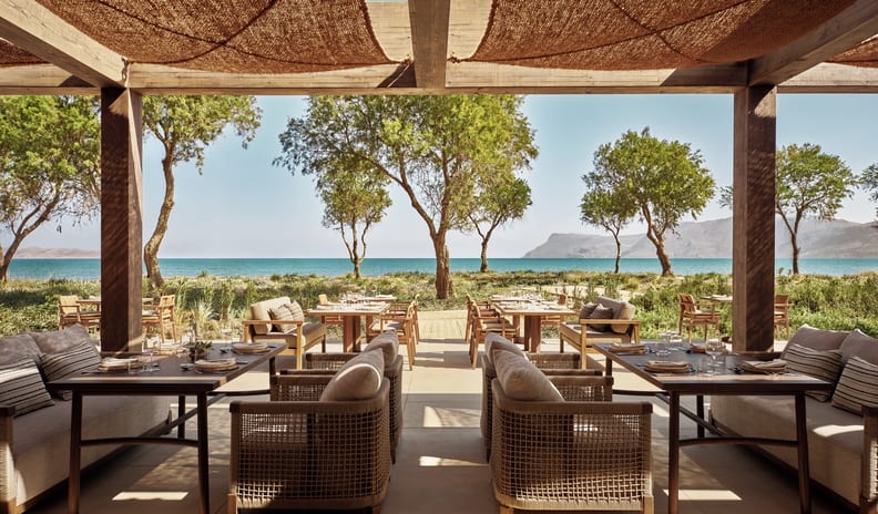 Beach Club Luxury Mediterranean style resort oceanfront outdoor dining patio under a woven pergola with wooden tables, lounge armchairs, and sea views at Ikos Kissamos by Kenwood Travel