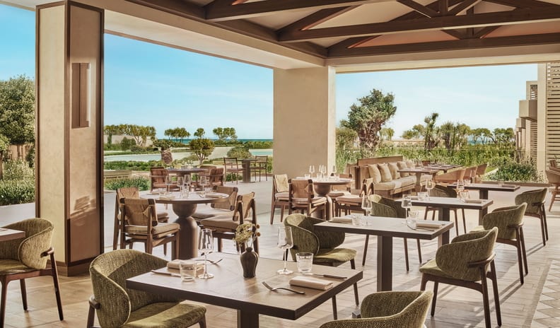 Fresco Luxury Mediterranean style resort covered patio restaurant featuring wooden beams, olive green dining chairs, and views of gardens and seaat Ikos Kissamos by Kenwood Travel
