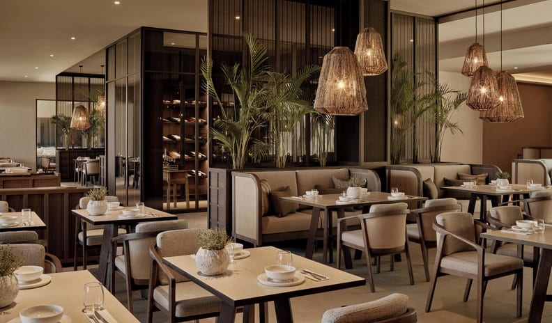 The Market Luxury Mediterranean style resort fine dining restaurant featuring plush booth seating, woven pendant lights, potted palms, and wine display cellar at Ikos Kissamos by Kenwood Travel