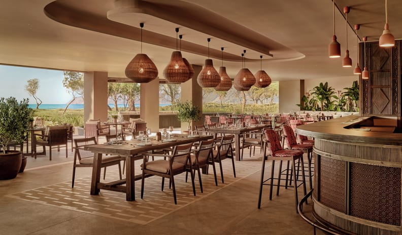 Azul Luxury Mediterranean style open-air resort restaurant and bar featuring woven pendant lights, wooden dining tables, bar seating, and coastal views at Ikos Kissamos by Kenwood Travel