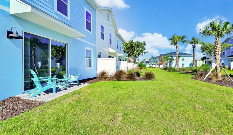 Four Bedroom Villa Back patio of a light blue coastal vacation home with turquoise Adirondack chairs, a small table, sliding glass door, and green lawn with palm trees at Margaritaville Resort Orlando by Kenwood Travel