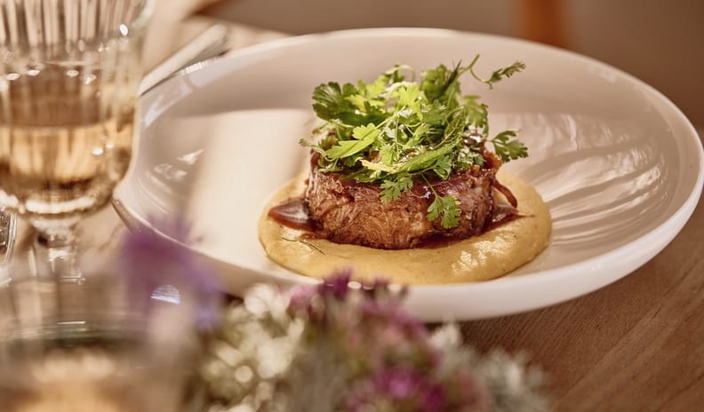 La Plage Gourmet braised beef dish served over a bed of creamy polenta, topped with fresh green herbs in a shallow white ceramic bowl at Ikos Kissamos by Kenwood Travel