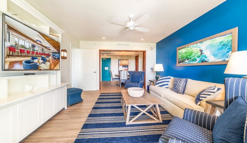One Bedroom Suite Spacious coastal living room suite featuring a cream sofa, vibrant blue accent wall with surf artwork, striped area rug, and custom entertainment center at Margaritaville Resort Orlando by Kenwood Travel