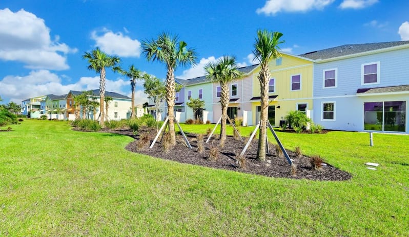 Three Bedroom Villa Row of colorful pastel resort townhomes featuring yellow, pink, and blue exteriors with a manicured green lawn and palm trees under a blue sky at Margaritaville Resort Orlando by Kenwood Travel