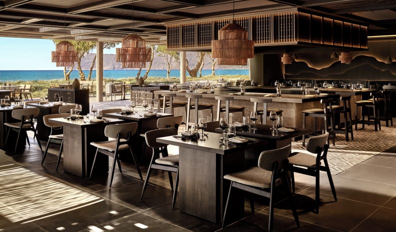Anaya Luxury oceanfront dining room featuring dark wood tables, grey upholstered chairs, large woven pendant lamps, and open views of the beach at Ikos Kissamos by Kenwood Travel
