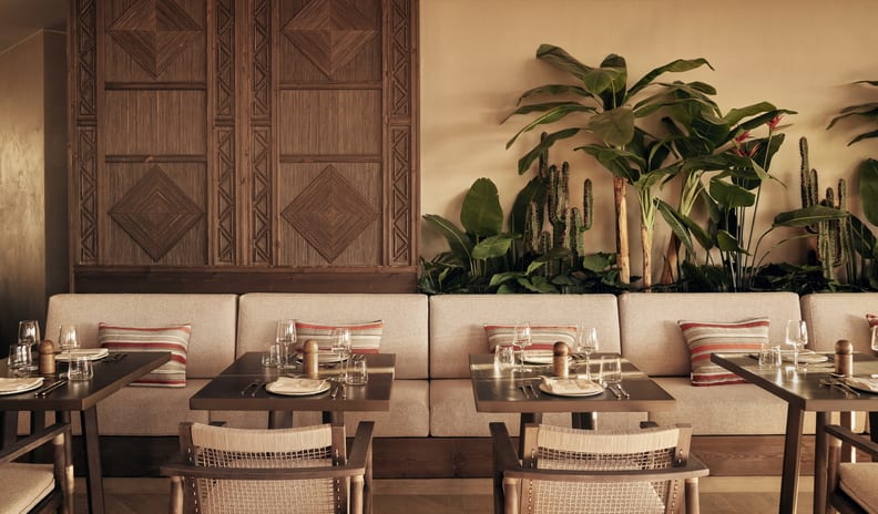 Azul Modern bohemian restaurant dining room featuring long upholstered banquet seating, wooden tables, striped accent pillows, and tropical potted plants against a warm beige wall at Ikos Kissamos by Kenwood Travel