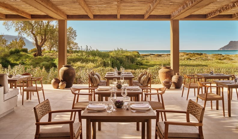 La Plage Open-air beachfront restaurant patio with wooden tables, woven armchairs, large ceramic terracotta urns, and panoramic ocean views under a thatched pergolas at Ikos Kissamos by Kenwood Travel