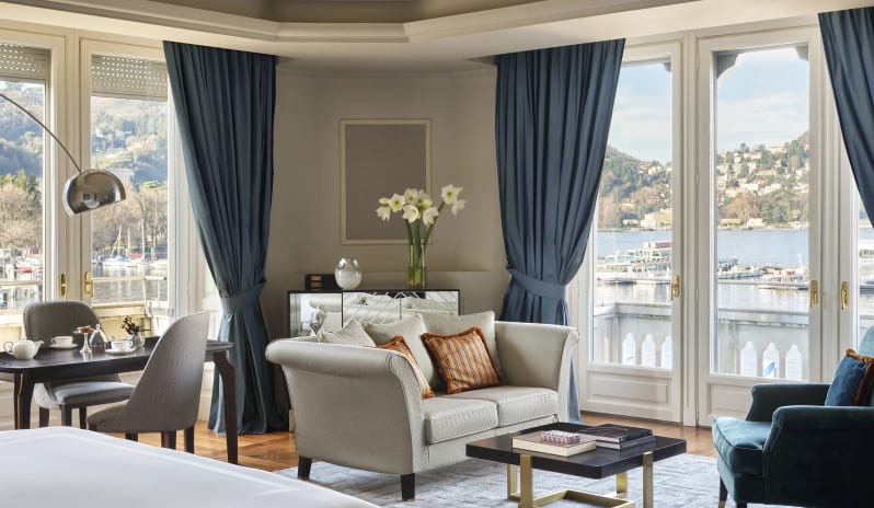 Corner Junior Suite living space with floor-to-ceiling lake-view windows and blue curtains at Palazzo Venezia by Kenwood Travel