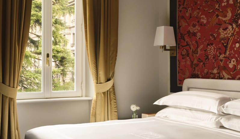 Junior Suite bedroom interior showcasing a red botanical wall panel, gold draped curtains, white pillows, and a bedside sconce lamp at Palazzo Venezia by Kenwood Travel