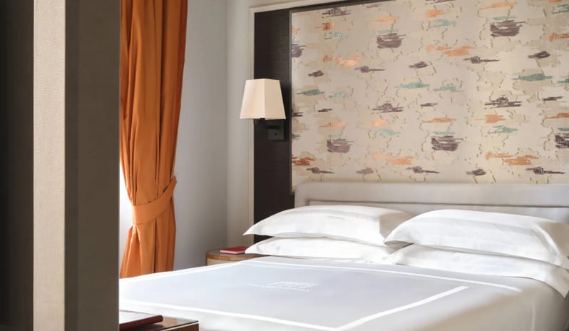 Deluxe Double Room bedroom with plush white bedding, decorative pattern headboard wall, wall sconce lamp, and rich terracotta-orange curtains at Palazzo Venezia by Kenwood Travel