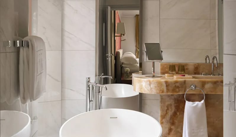 Junior Suite bathroom with a round white soaking tub, chrome fixtures, carved amber stone vanity sink, and bright white marble wall tiles at Palazzo Venezia by Kenwood Travel