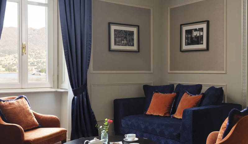 Suite seating area with terracotta velvet armchairs, navy sofa, dark round coffee table with tea set, and parquet floors by a lake-view window at Palazzo Venezia by Kenwood Travel